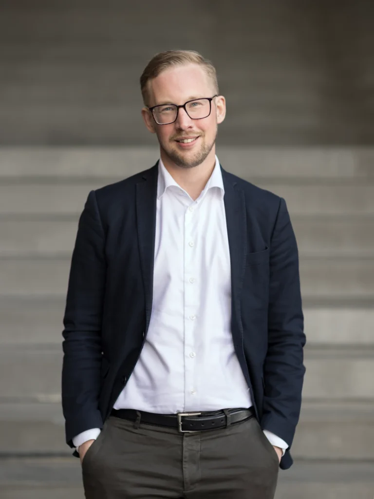 En man med kort blont hår och glasögon, klädd i mörk kavaj, vit skjorta och grå byxor, står med händerna i fickorna och ler framför en suddig betongtrappa.