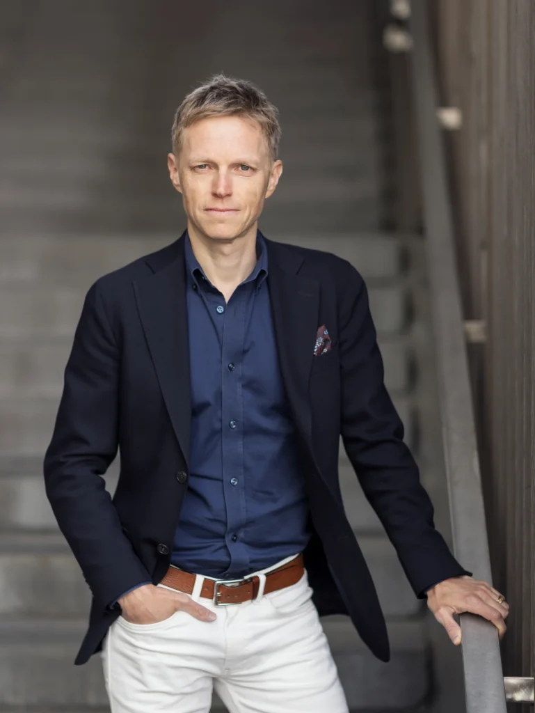 En man med kort blont hår, klädd i marinblå kavaj, blå skjorta, vita byxor och brunt bälte, står med ena handen i fickan och den andra vilande på ett räcke vid en trappa.